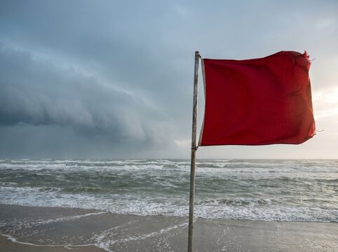 Red warning flag on a beach with stormy sky and rough ocean waves, indicating hazardous conditions.