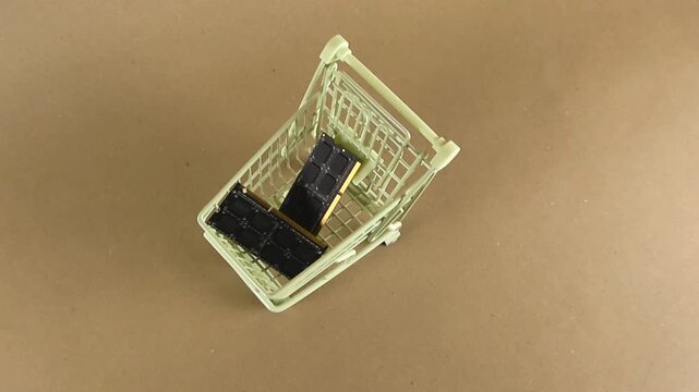 Small shopping cart carrying two computer RAM modules spinning on a brown background, symbolizing the purchase of PC components, hardware upgrades, and the high cost of technology
