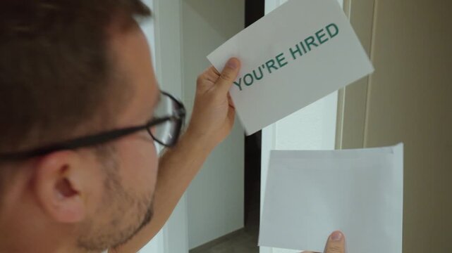 Man reveals a card reading &ldquo;YOU&rsquo;RE HIRED&rdquo; and celebrates with a fist gesture, representing a successful job application, recruitment and new employment.