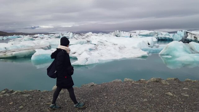A female traveler in a black parka and scarf walks along the pebble shore of Jokulsarlon as white and blue icebergs float in the calm turquoise lagoon.