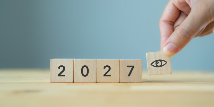 2027 Outlook, Future Vision and Strategic Planning.  Hand placing an eye symbol wooden block beside 2027 number. Future outlook, vision, forecasting and strategic planning against a blue background.