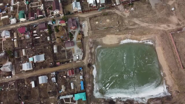 Drone hovers above rural homes near frozen pond. Green water churns beneath icy surface. Snow lines edge of frozen pool. Villagers live nearby, quiet, unbothered