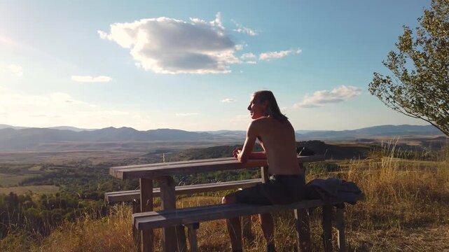 Shirtless man sitting on a wooden bench looking at the mountain view 