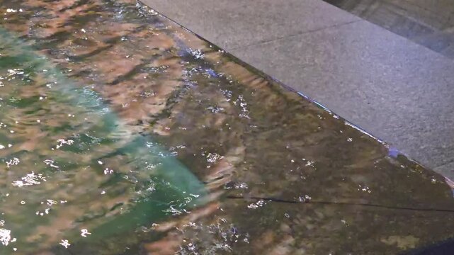 Close up of clear water flowing smoothly over the polished stone edge of a modern fountain pool creating gentle ripples and sparkling reflections
