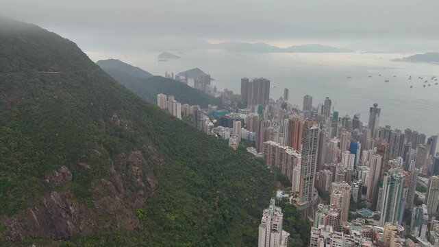 Aerial flight along the green forested slope of Victoria Peak above the Mid-Levels residential towers of Hong Kong Island, grey sea haze and outlying islands in the distance