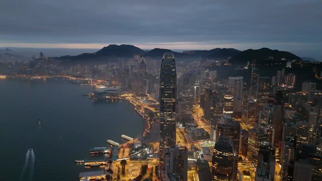 Forward drone flight over Wan Chai toward the tower and Central at blue hour, glowing harbourfront roads along Victoria Harbour and lit homes on Victoria Peak