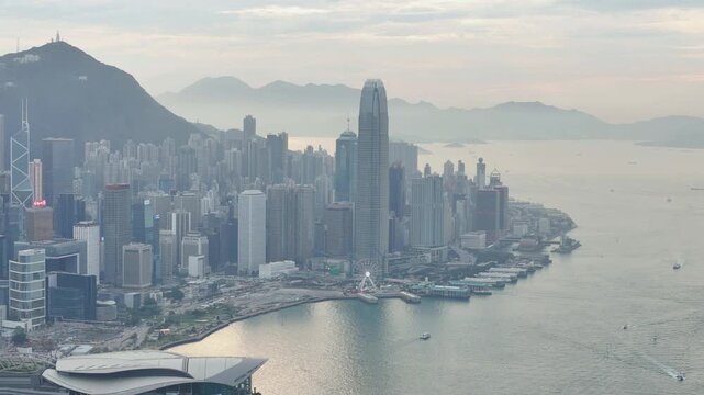 Central and Wan Chai skyline beneath Victoria Peak in soft hazy light, slow aerial pull back revealing the tower and the glittering harbourfront of Hong Kong Island