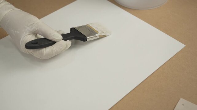 Gloved hand using a paintbrush to apply white paint or primer to a sheet of paper or board