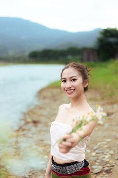 Young Asian woman holding wildflower bouquet beside tranquil mountain lake, smiling outdoors with natural joy and scenic countryside surroundings