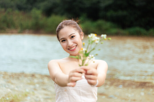 Smiling young Asian woman holding delicate white flower bouquet beside calm lake, creating joyful outdoor portrait with soft natural light and blurred woodland background
