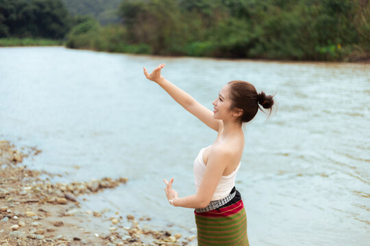 Young Asian woman practicing meditation beside calm river, raising arms with peaceful focus near rocky shore and green forest