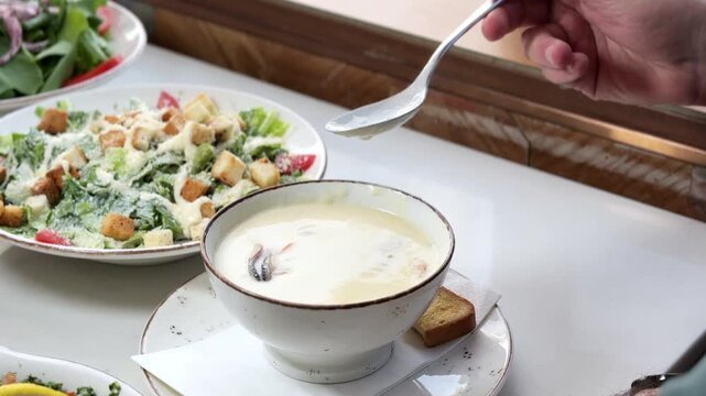 A close up of a person using a spoon to eat creamy seafood soup at a bright indoor restaurant. The delicious hot meal is served on a white table alongside a fresh caesar salad for lunch.