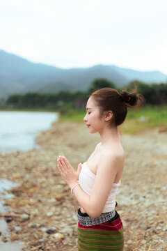 Young Asian woman in traditional clothing praying beside mountain lake on quiet rocky shore, with peaceful expression, green landscape, and soft natural light creating serene scene