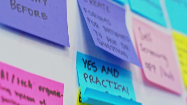 Close-up of handwritten sticky notes in pink, yellow, blue and purple stuck to a white wall during a brainstorming session. The camera pans across the notes with shallow depth of field, showing ideas