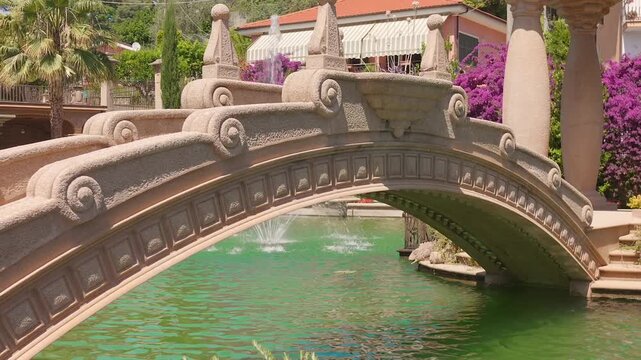 Decorative Villa Grock arch footbridge spans a green pond with fountains and lush flowers in the garden sun.