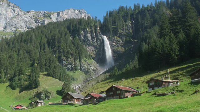 Waterfall near Asch in Uri, Switzerland. Waterfall above wooden chalets in Unterschachen. Staubifall waterfall surrounded by alpine forest. Swiss waterfall beneath rugged mountain cliffs.