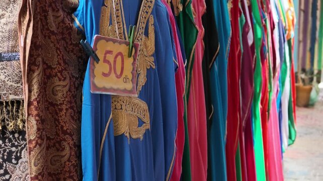 Close-up panning shot showcasing a vibrant rack of colorful traditional garments, fabrics, and textiles hanging outside a local shop in Fes