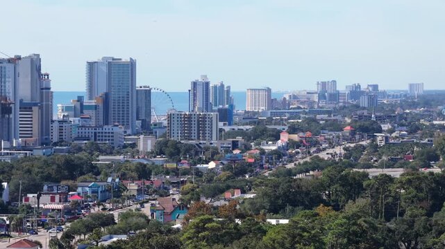 Cars and colorful shops line a busy road toward a skyline of high rise hotels with a Ferris wheel and the ocean beyond in Myrtle Beach, South Carolina, USA. Aerial approaching shot