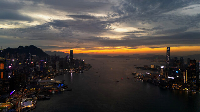 Aerial drone view of Hong Kong at twilight, a golden strip of afterglow above the outer islands with Victoria Harbour glowing between the island and Kowloon skylines