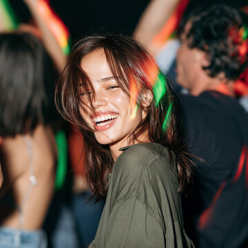 A young woman laughs gleefully while dancing in a nightclub, illuminated by colorful lights. Her eyes are closed and her head is tilted back in pure enjoyment