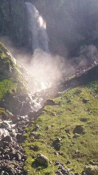 Waterfall closeup with falling water and mist. Waterfall detail against dark textured rock. Vertical waterfall view with sunlit spray. Scenic waterfall background with rugged cliff walls.