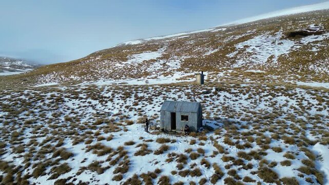 Nicholsons Hut sits in a remote snowy New Zealand range as two people linger outside while the drone pulls back to reveal the empty landscape.