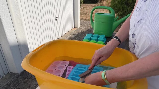Woman washes colorful silicone molds outdoors in a tub filled with water. She scrubs one mold with a dish brush, turns it over to clean the other side and places it back into the water. Static shot