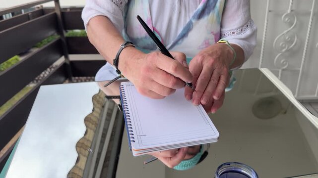 Woman sits at a table on a balcony and tests a freshly filled fountain pen by writing in a spiral notebook. Static shot.