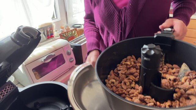Woman opens an air fryer and pours freshly roasted mixed nuts into a stainless steel bowl. Almonds, cashews and walnut halves are visible in the homemade snack mixture, Static shot.