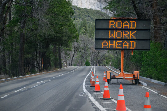 A portable electronic message sign on a trailer displaying "ROAD WORK AHEAD", accompanied by orange traffic cones along the road inside Yosemite National Park. California.