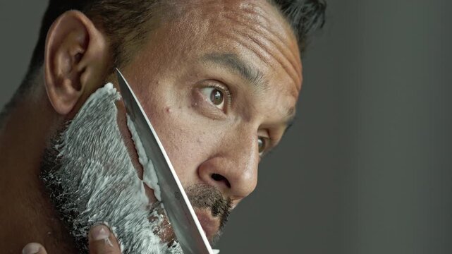 Man shaving his cheek with a knife. Man with shaving cream covering his beard. Closeup of man holding a sharp blade. Bearded man during an unconventional shaving routine.