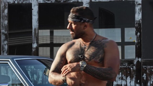 Gangster fashion with jewelry and chest tattoos. Gangster aesthetic with a patterned headband. Shirtless man in gangster style closeup. Urban gangster portrait against a blurred background.