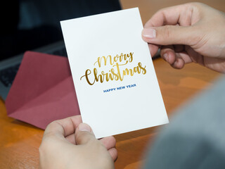Person holding Merry Christmas and Happy New Year greeting card with red envelope at work desk. Corporate holiday communication, client appreciation, seasonal wishes and business greetings concepts. © tete_escape