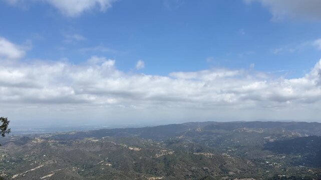 A wide static view of rolling ridges and canyons of the Santa Monica Mountains from Lois Ewen Overlook in Topanga, California, under a bright blue sky with a long band of white clouds.