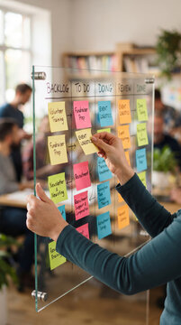 Colleague Placing Sticky Notes on Transparent Glass Kanban Board in Office