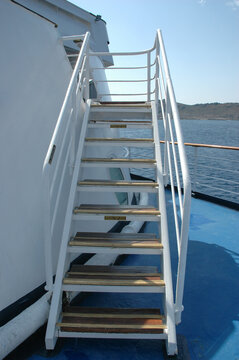 A straight on view of white metal stairs with wooden steps on a blue ship deck leading to an upper level under a blue sky.