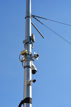A detailed low angle view shows multiple surveillance dome and bullet cameras with electronic equipment attached to a metal utility post against a blue sky.