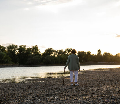 A senior woman stands on a river shore with a cane at evening