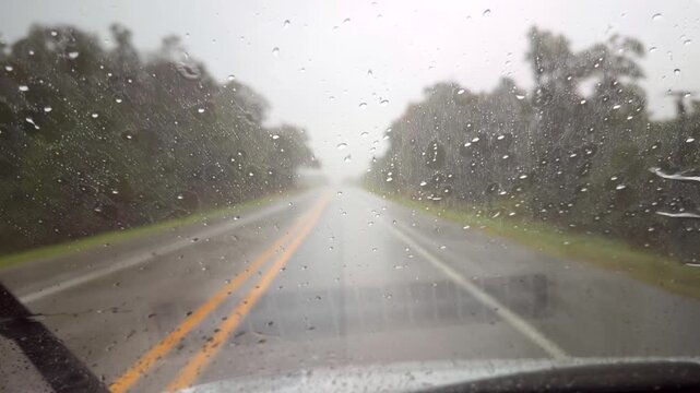 Heavy rain pelts a car windshield as wipers sweep across a wet, empty rural highway, creating a tense driver&rsquo;s-eye view in reduced visibility.