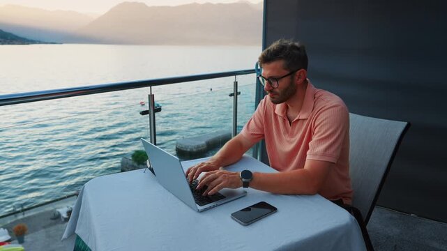 Focused remote worker typing on laptop at balcony table overlooking serene lake and mountains during peaceful sunset, balancing productivity with tranquil outdoor view