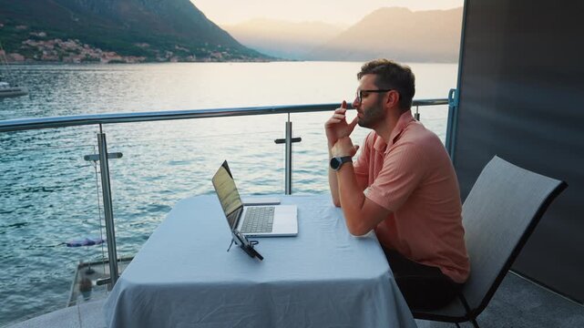 Remote professional working on laptop during peaceful waterfront sunset, focused on online communication from scenic balcony overlooking calm coastal mountains