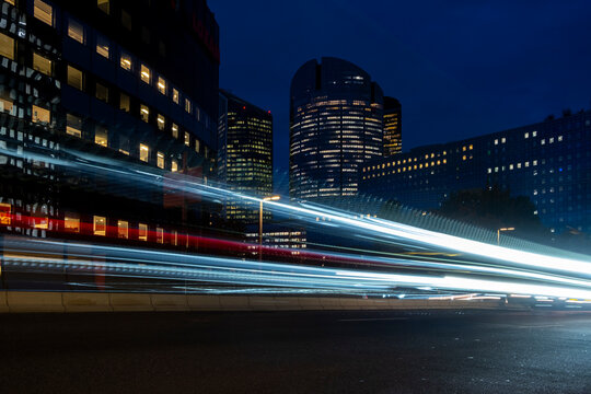 Light trails sweep through a night city beside office buildings as traffic motion crosses the street and the trails continue between the buildings and architecture