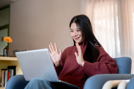 Young Asian woman waving hands, gesturing on laptop call on sofa in a living room or cafe.