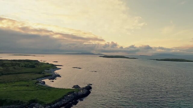 Renvyle coast in Connemara, Ireland, shows a rocky green shoreline and calm sea, with low offshore islands, distant hills and warm golden hour light.