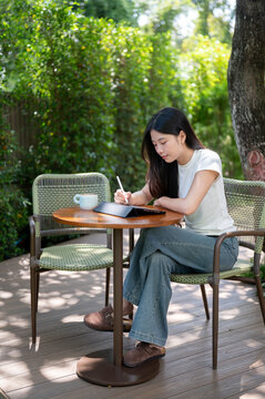 Asian Woman Writing on Tablet with Stylus at Garden Patio Table