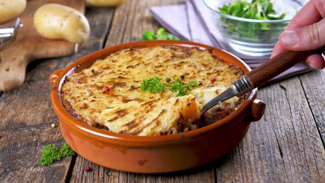 hand serving cottage pie with spoon from ceramic dish on wooden background