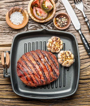 Grilled beef ribeye steak in cast iron pan with garlic and seasonings on wooden table