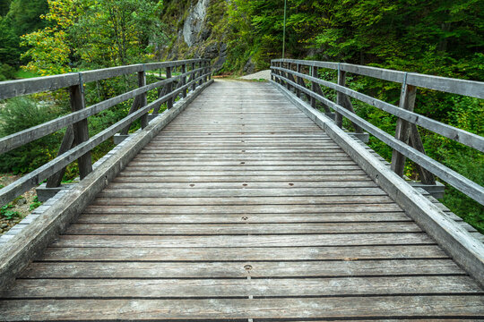 Wide wooden plank bridge with timber guardrails leading over mountain river gorge into lush green forest.