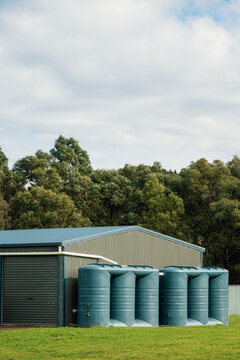 Two large slimline polyethylene rain water tanks alongside a steel shed on a farm set against a backdrop of dense eucalyptus trees under a broad, cloud-filled sky, Victoria, Australia