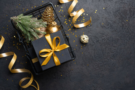 Black gift box with gold bow and Christmas decorations in a wire basket on dark grunge background